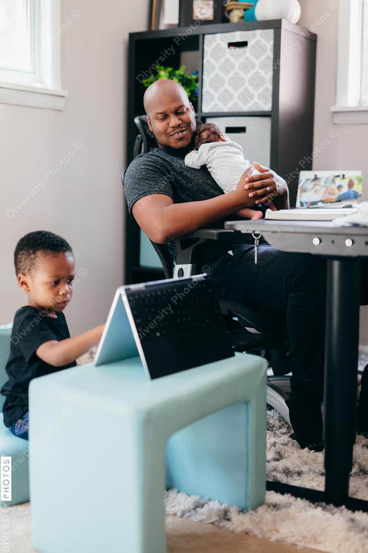 Black dad with newborn working from home helping son with schoolwork 