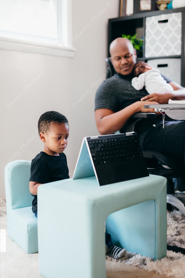 Black dad with newborn working from home helping son with schoolwork 