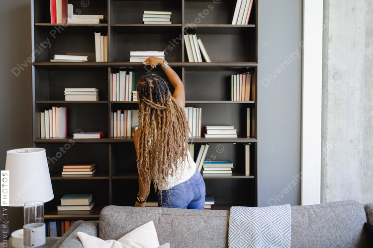 Rear view of woman with long dreadlocks reaching for book in living room