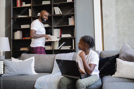 Black Boy using laptop on sofa in living room, father at bookshelf in background