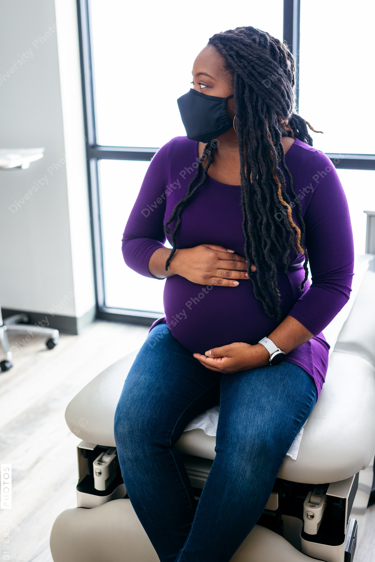 Black pregnant woman waiting in medical exam room at the Doctors office, wearing mask
