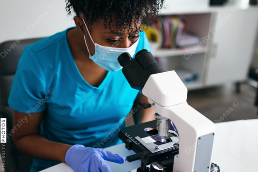 Black medical technologist in mask in laboratory looking through microscope at blood samples