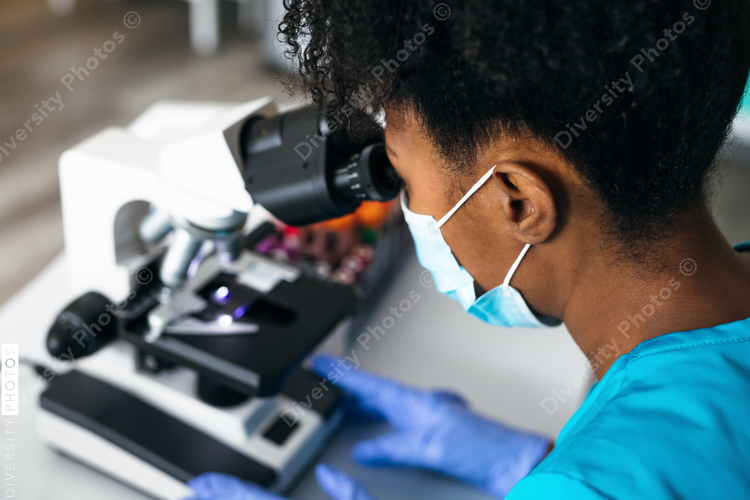 Black medical technologist in mask in laboratory looking through microscope at blood samples