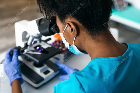 Black medical technologist in mask in laboratory looking through microscope at blood samples