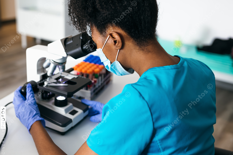 Black medical technologist in mask in laboratory looking through microscope at blood samples