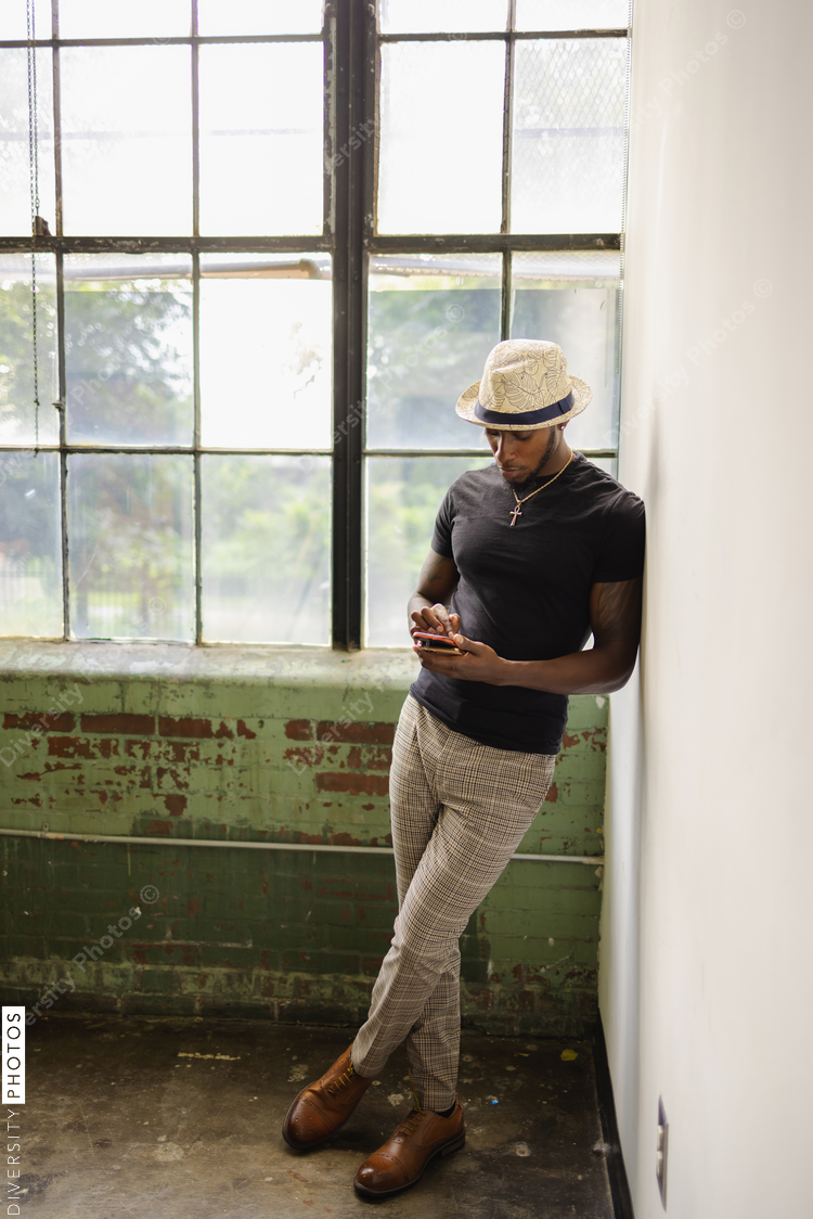 Elegant young Black man standing next to window and using phone, fashionable