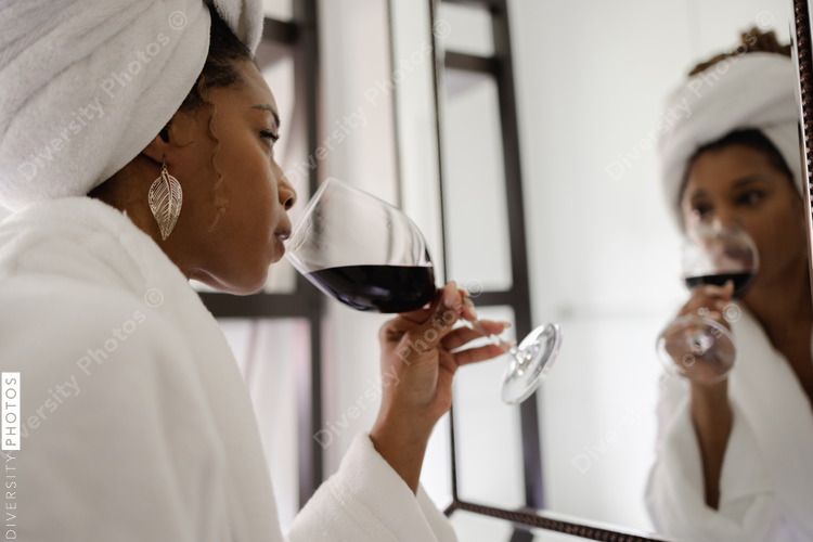 Woman in bathrobe looking in bathroom mirror while drinking wine