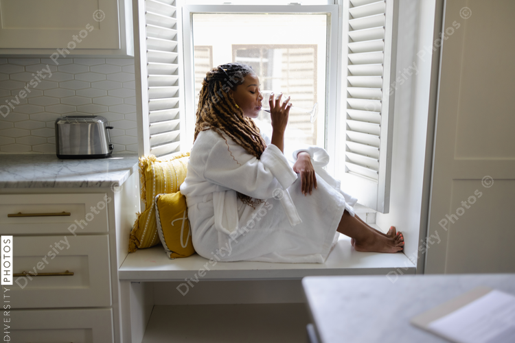 African American Woman in bathrobe sitting by window, drinking wine, cozy