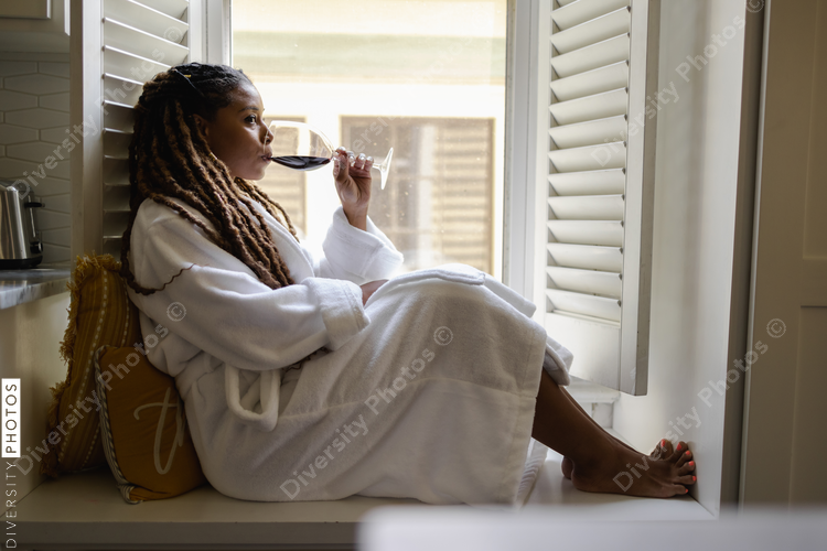 Woman in bathrobe sitting by window, drinking wine