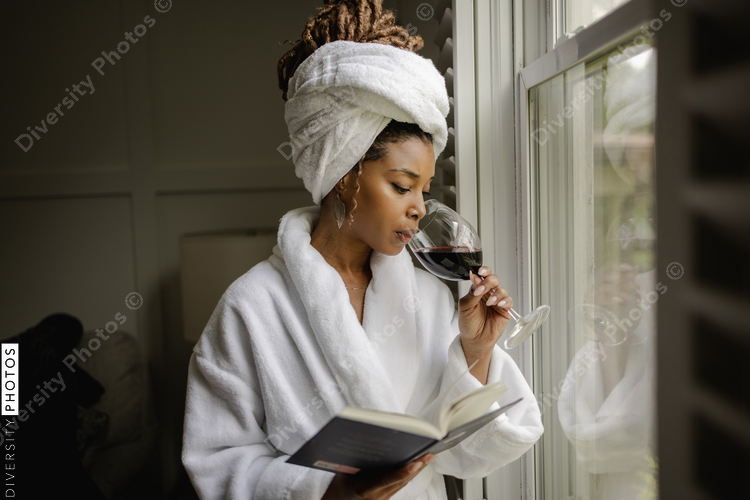 Woman in bathrobe standing by window, holding book and glass of wine