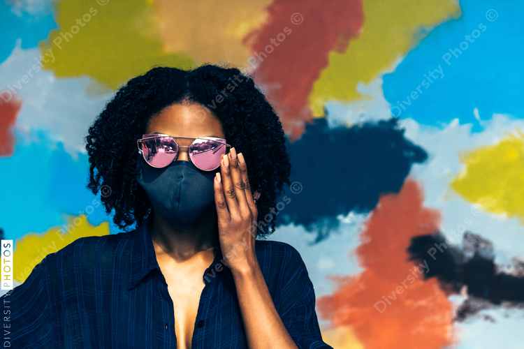 Black woman with natural hair wearing mask and sunglasses in front of mural