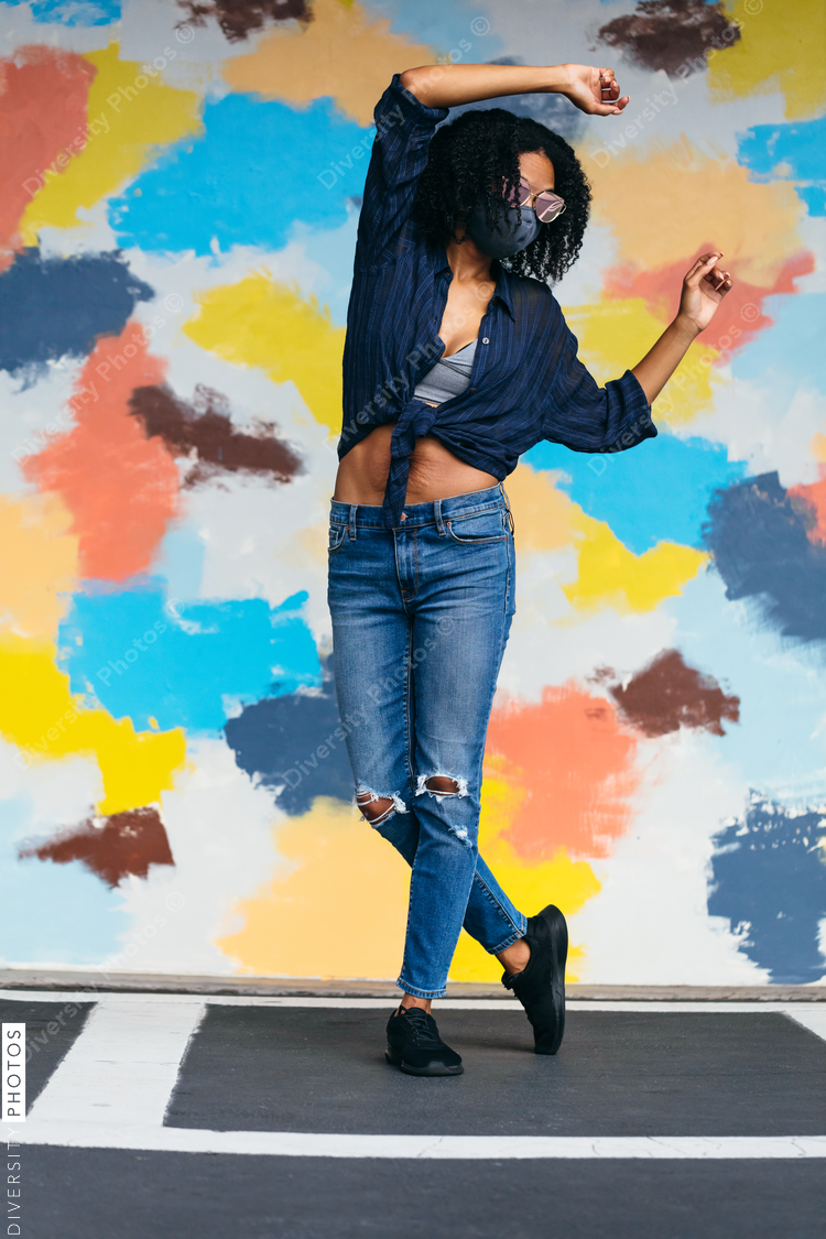 Black woman with natural hair wearing mask and sunglasses dancing in front of mural