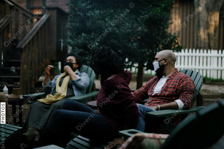 Black friends sitting around firepit safely wearing mask in backyard
