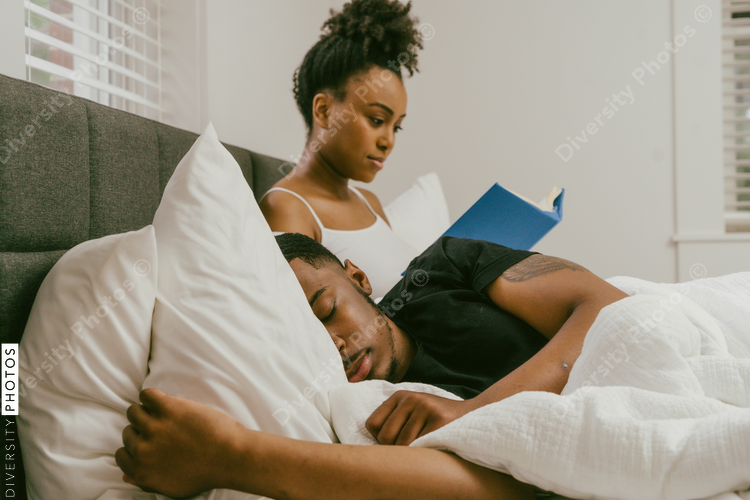 Couple laying in bed at home, boyfriend sleeping and girlfriend reading a book