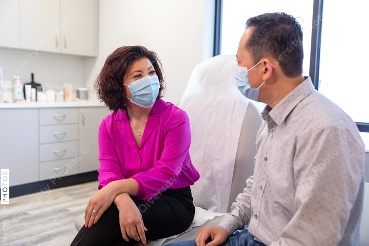 Middle aged Asian couple waits for doctor in medical exam room wearing mask