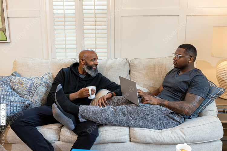 Black same-sex couple on couch, cuddled and working on laptop at home