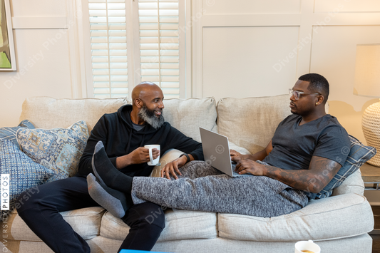 Black same-sex couple on couch, cuddled and working on laptop at home