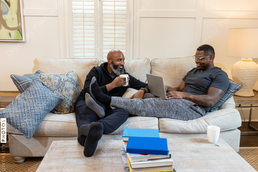 Black same-sex couple on couch, cuddled and working on laptop at home