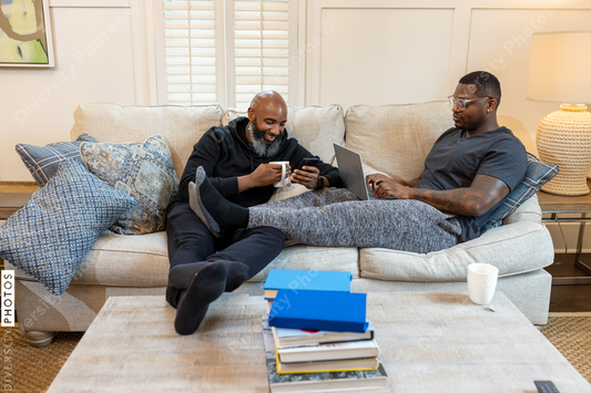 Black same-sex couple on couch, cuddled and working on laptop at home