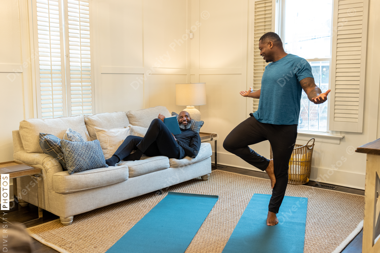 Man encourages partner on couch while he does yoga at home, fitness
